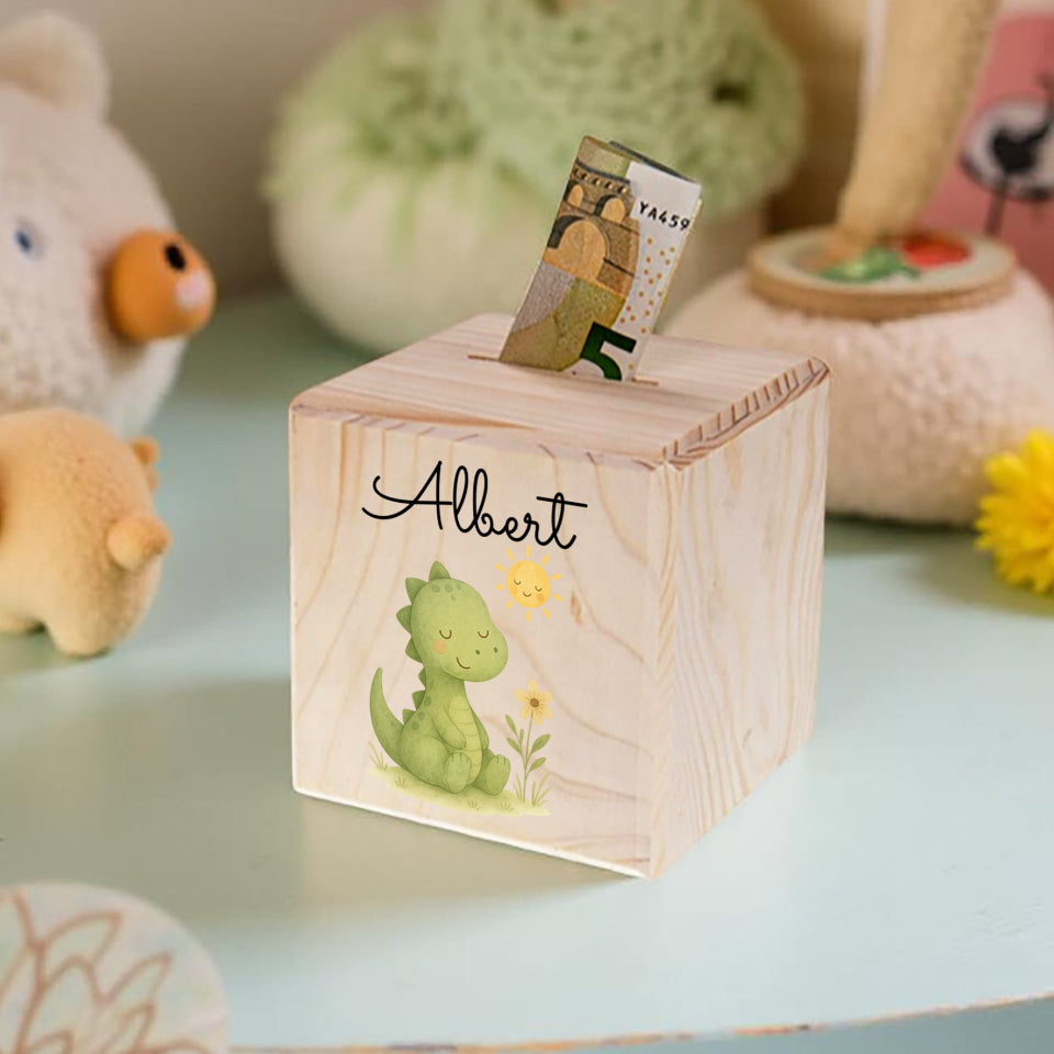 Wooden money box with a dinosaur design and the name 'Albert' on a light blue surface with toys in the background.