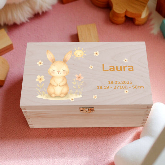 Wooden box with a rabbit design and 'Laura' text, surrounded by wooden toys and plants on a pink surface.