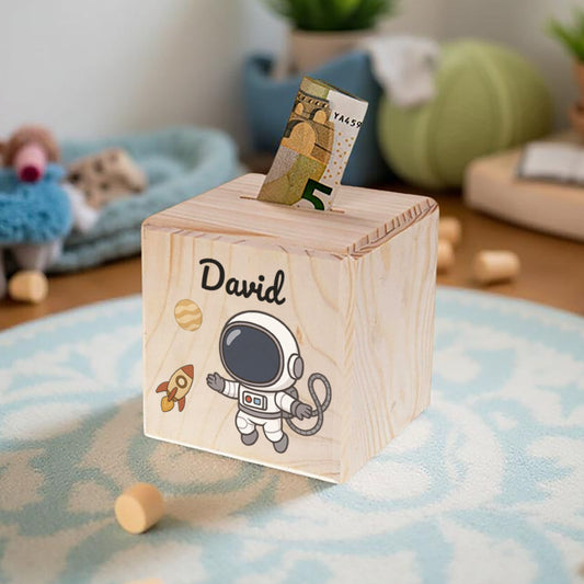 Wooden money box with 'David' and an astronaut design, placed on a surface with toys and plants in the background.