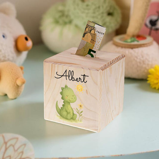 Wooden money box with a dinosaur design and the name 'Albert' on a light blue surface with toys in the background.