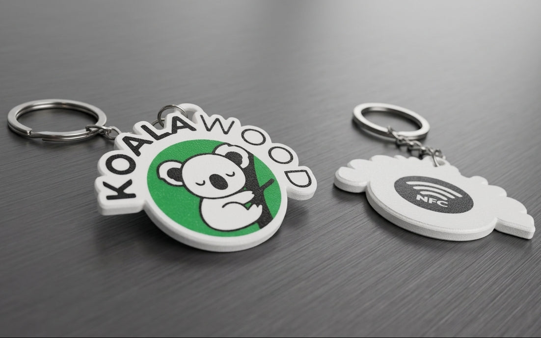 Two custom rubber keychains on a dark gray wood-grain surface. One keychain features a green circular charm with a cartoon panda and the text 'KOALAWOOD' in white.