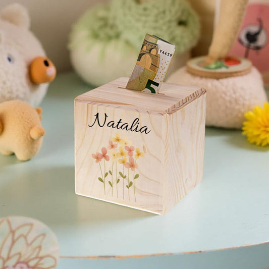 Wooden money box with floral design and 'Natalia' engraving on a light blue surface with toys and a flower in the background.