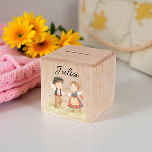 Wooden money box with 'Julia' and illustrations of a boy and girl on a white surface with yellow flowers in the background.