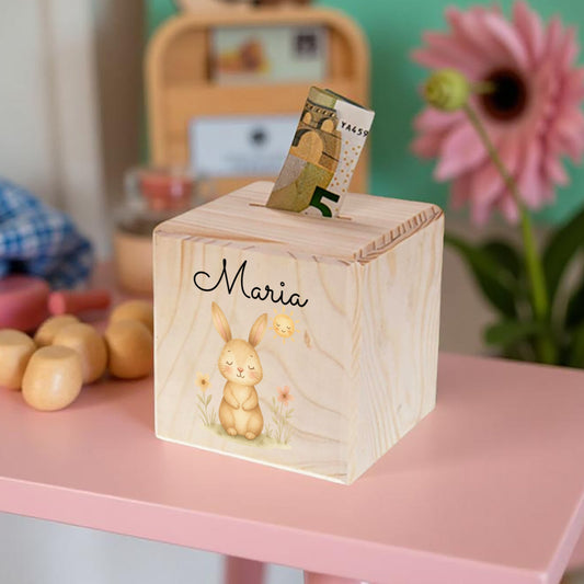 Wooden money box with a rabbit design and the name 'Maria' on a pink surface.