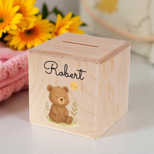 Wooden money box with a teddy bear illustration and 'Robert' engraving, placed on a surface with flowers and a pink blanket in the background.
