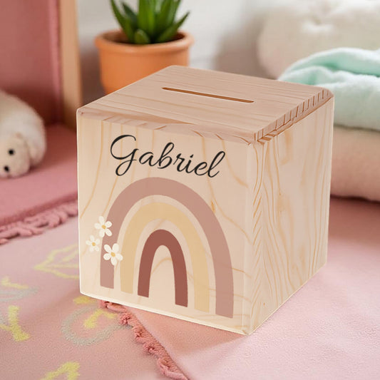 Wooden money box with rainbow design and 'Gabriel' engraving on a pink surface.