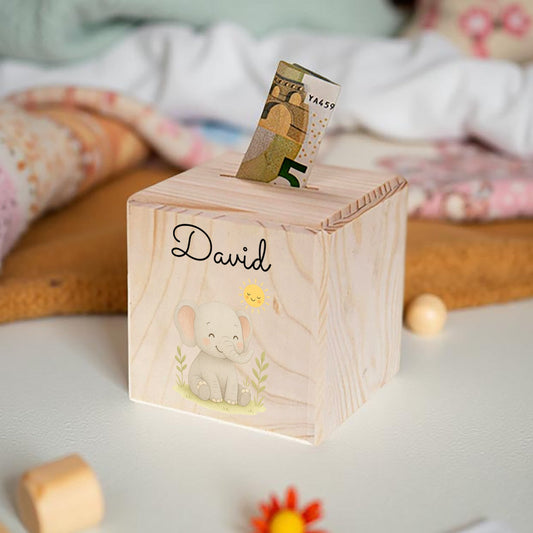 Wooden money box with a name engraving on a bed with colorful pillows and blankets.