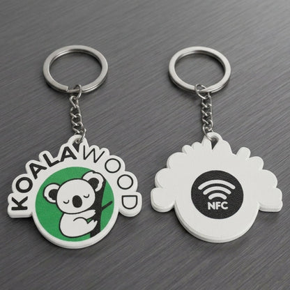 Two custom keychains on a dark gray wood-textured background. One keychain has a green and white koala logo with the text 'KOALAWOOD'.